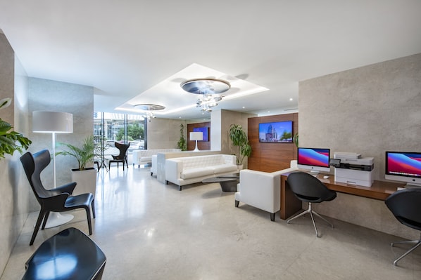 Un área de recepción moderna con un sofá blanco, sillas negras, un mostrador con computadoras y un televisor grande en la pared.