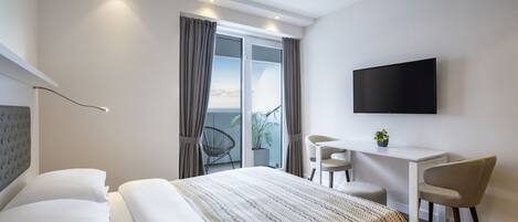 A modern hotel room with a large bed, a desk, a chair, a TV, and a balcony with curtains.