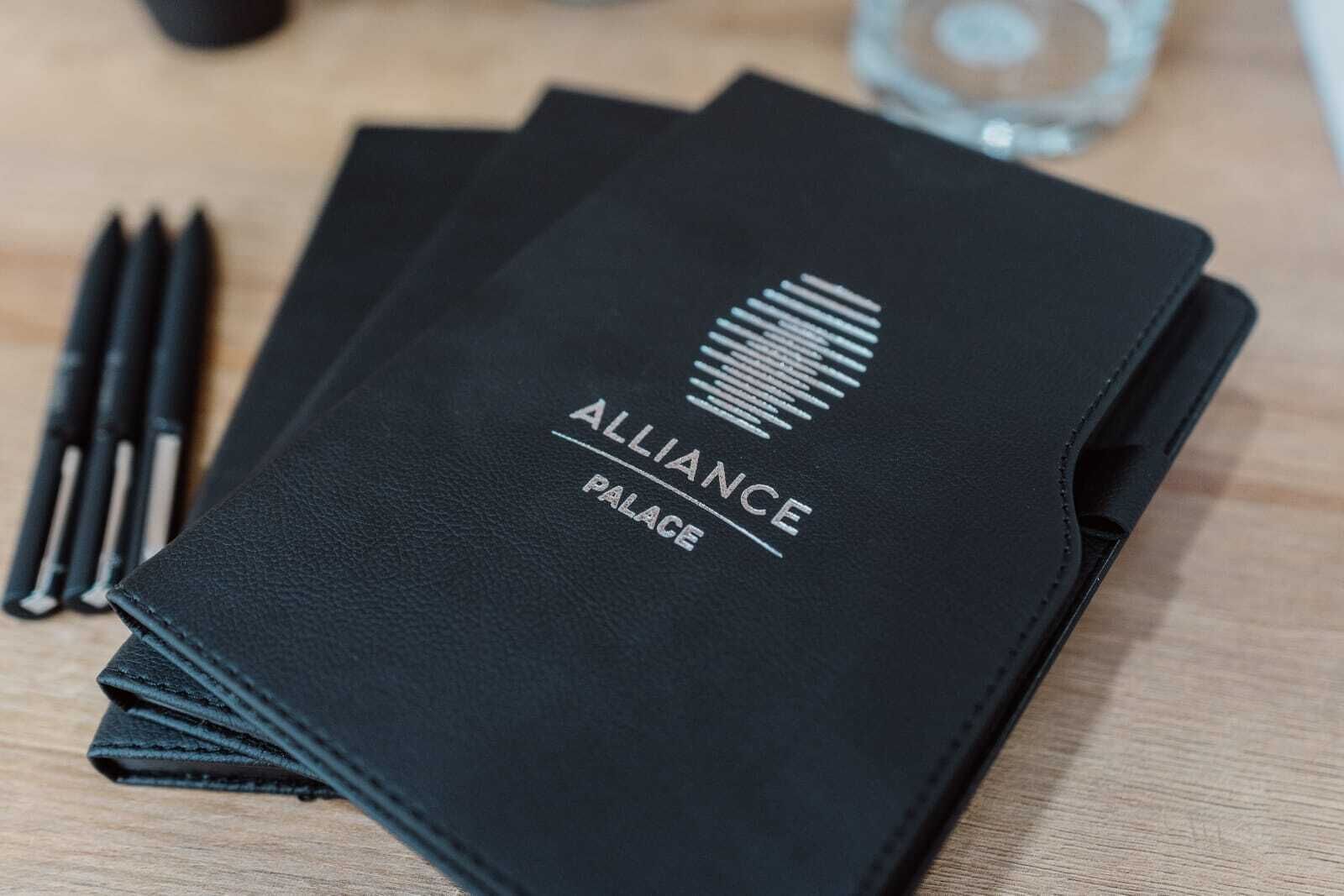 A black leather folder with the text "ALLIANCE PALACE" and a logo, placed on a wooden surface with pens nearby.