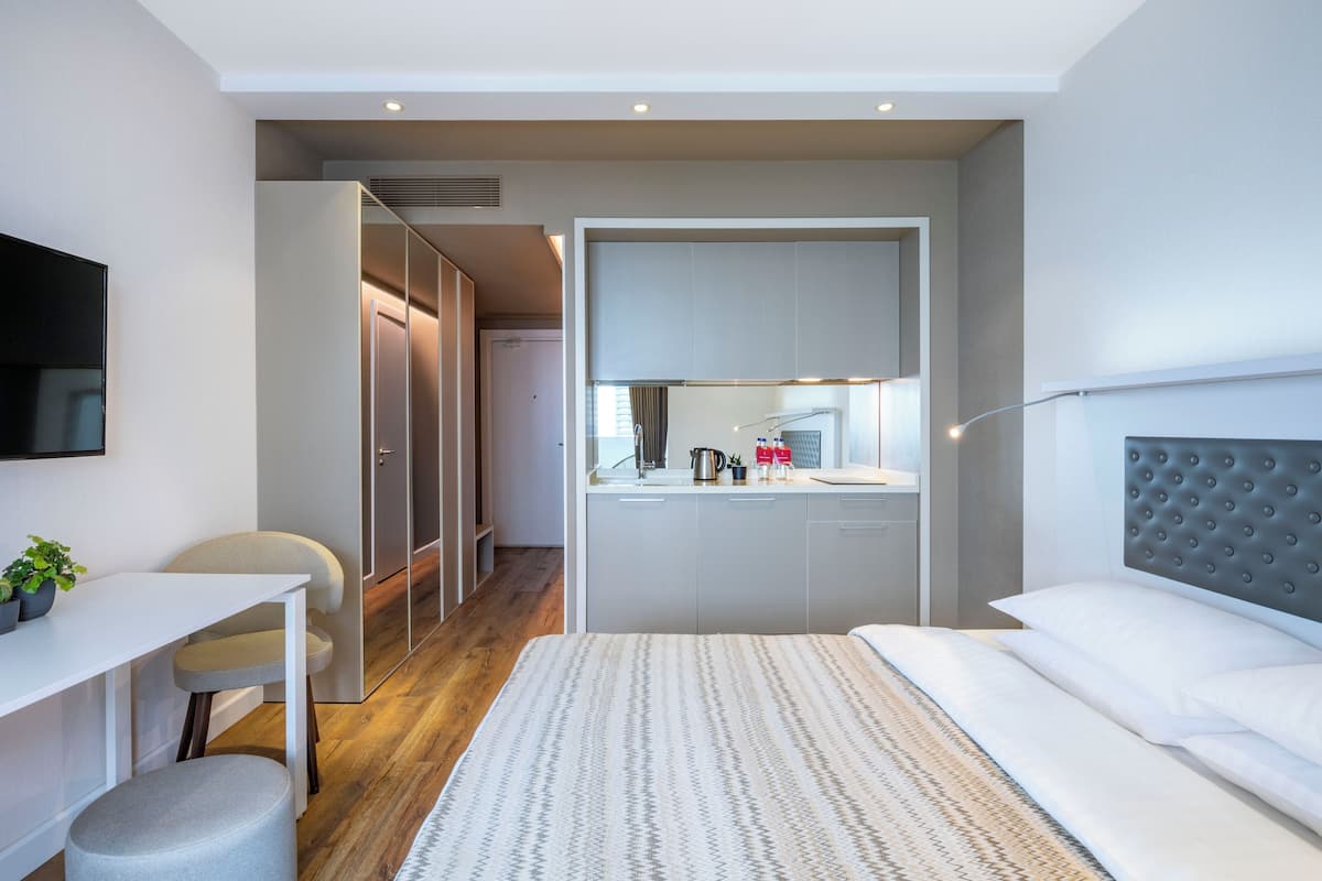 A modern hotel room with a bed, a desk, a chair, and a kitchenette.