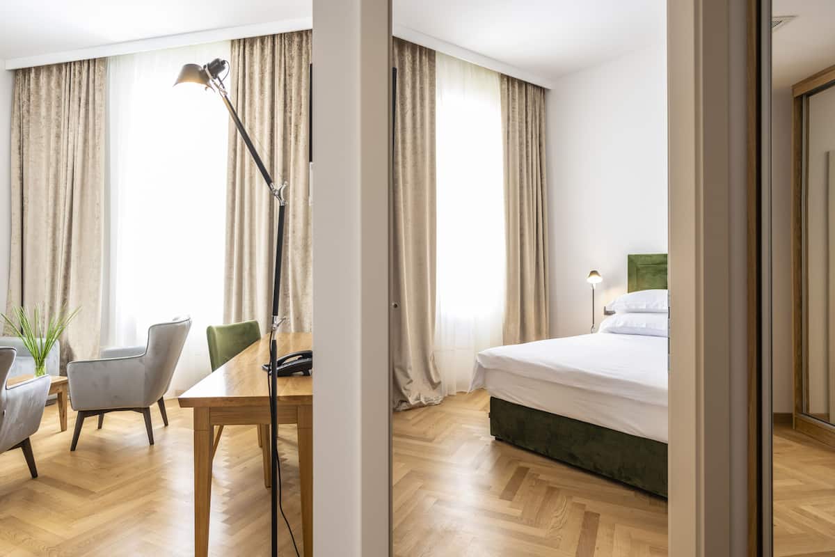 A hotel room with a wooden floor, a bed with a green headboard, a desk with a telephone, and a chair.