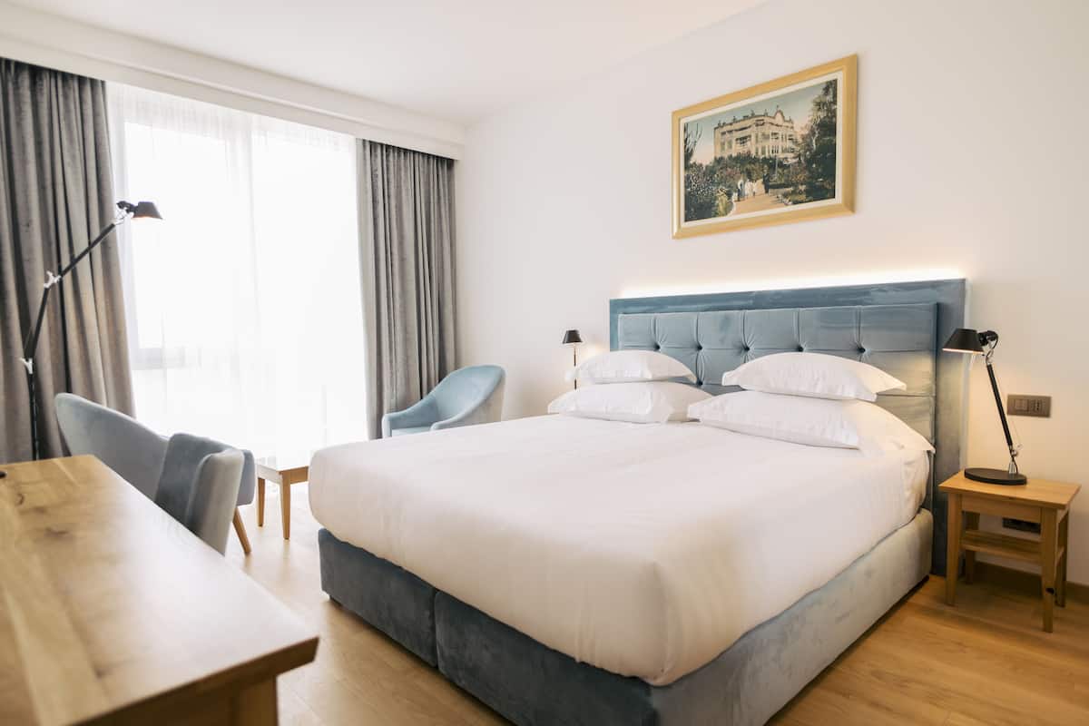 A hotel room with a large bed, a desk, a chair, a lamp, and a framed picture on the wall.