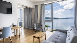 Un salon moderne comprenant un canapé, une table basse, un bureau avec une chaise, un téléviseur et une grande fenêtre donnant sur la mer.