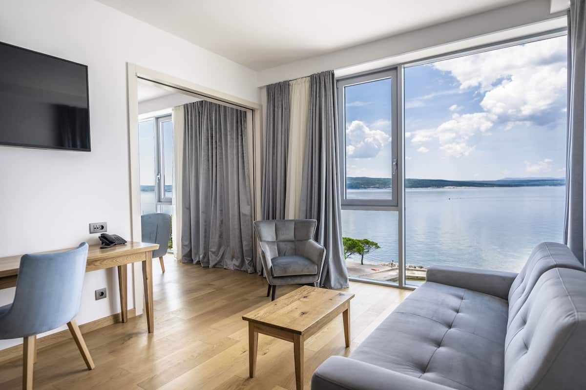 A modern living room with a sofa, a coffee table, a desk with a chair, a television, and a large window with a view of the sea.