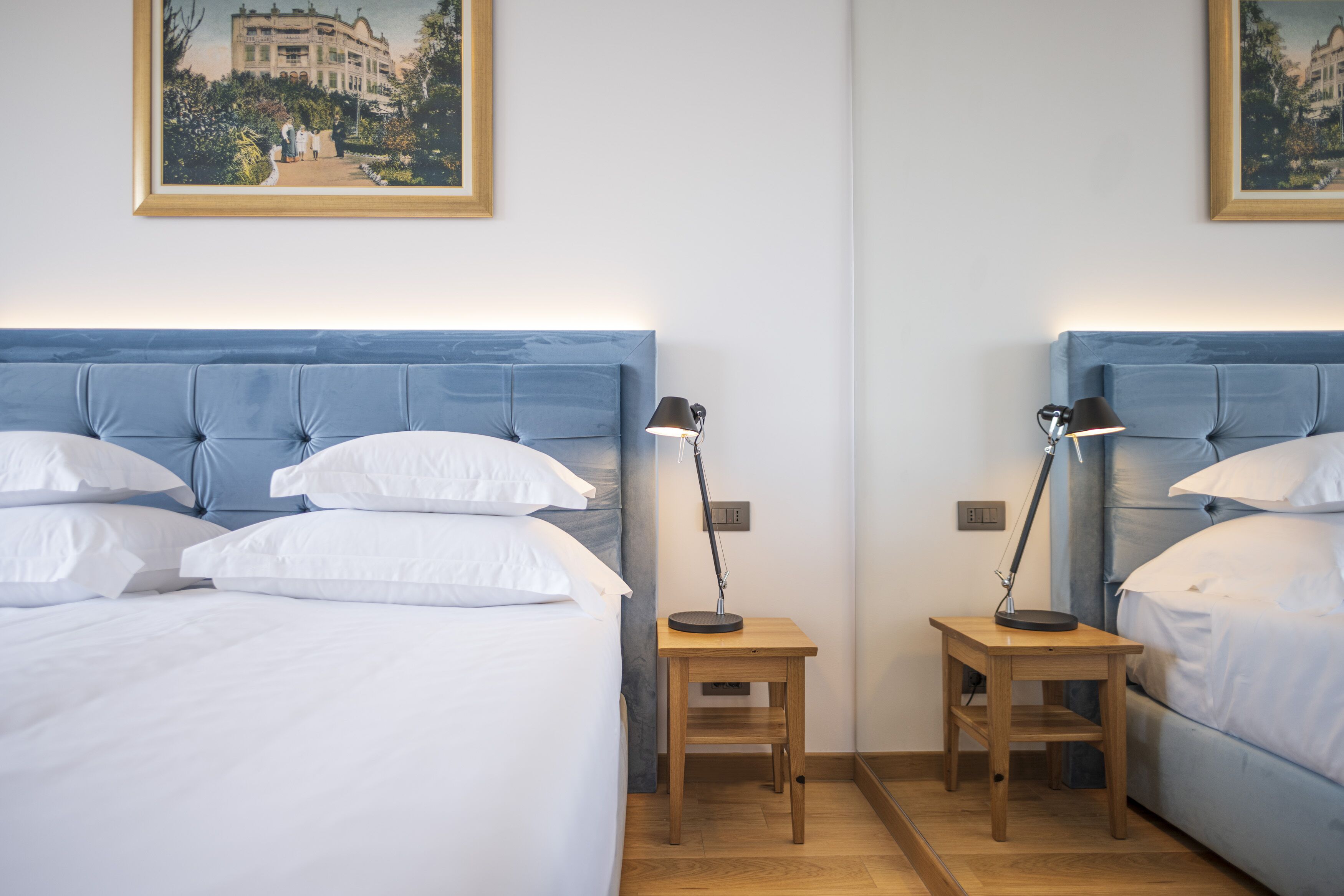 A hotel room with two beds, each with a blue headboard, white linens, and a small wooden nightstand with a lamp.