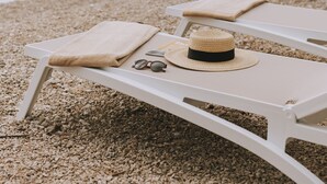 Une scène de plage avec deux chaises longues blanches, un chapeau de paille et une paire de lunettes de soleil posées sur l’une d’elles.