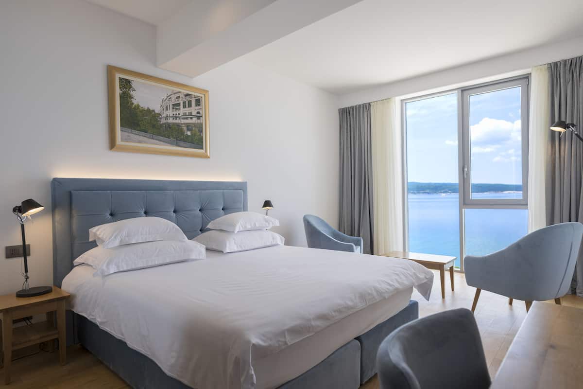 A modern hotel room with a large bed, a desk, two chairs, and a large window offering a view of the sea.