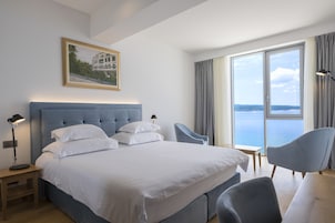 Moderni hotellihuone, jossa on suuri sänky, työpöytä, kaksi tuolia ja suuri ikkuna, josta avautuu näkymä merelle.