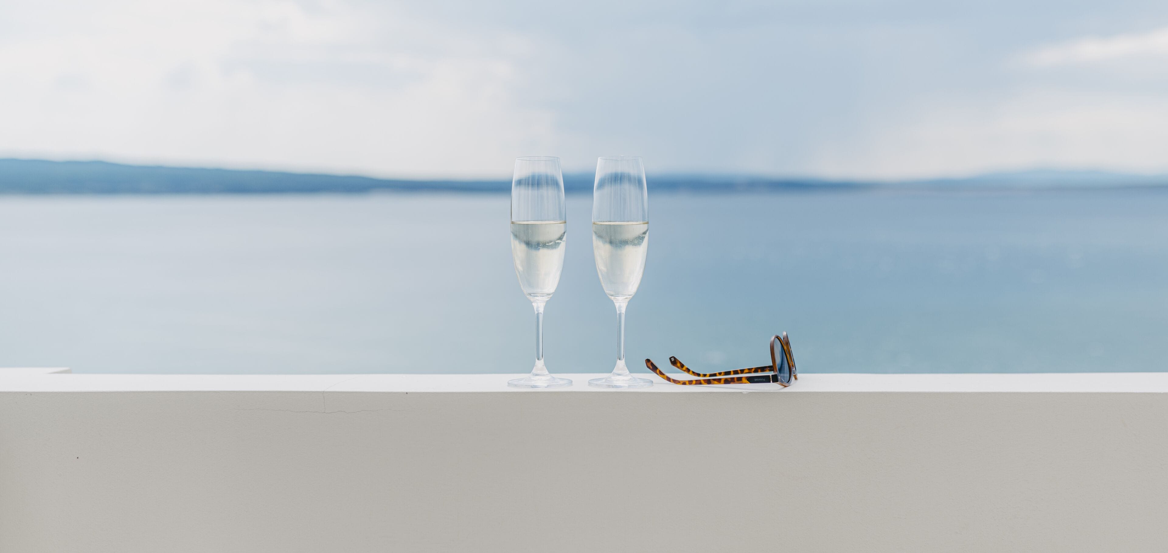 Two glasses of water on a ledge with a pair of sunglasses beside them, overlooking a calm body of water.