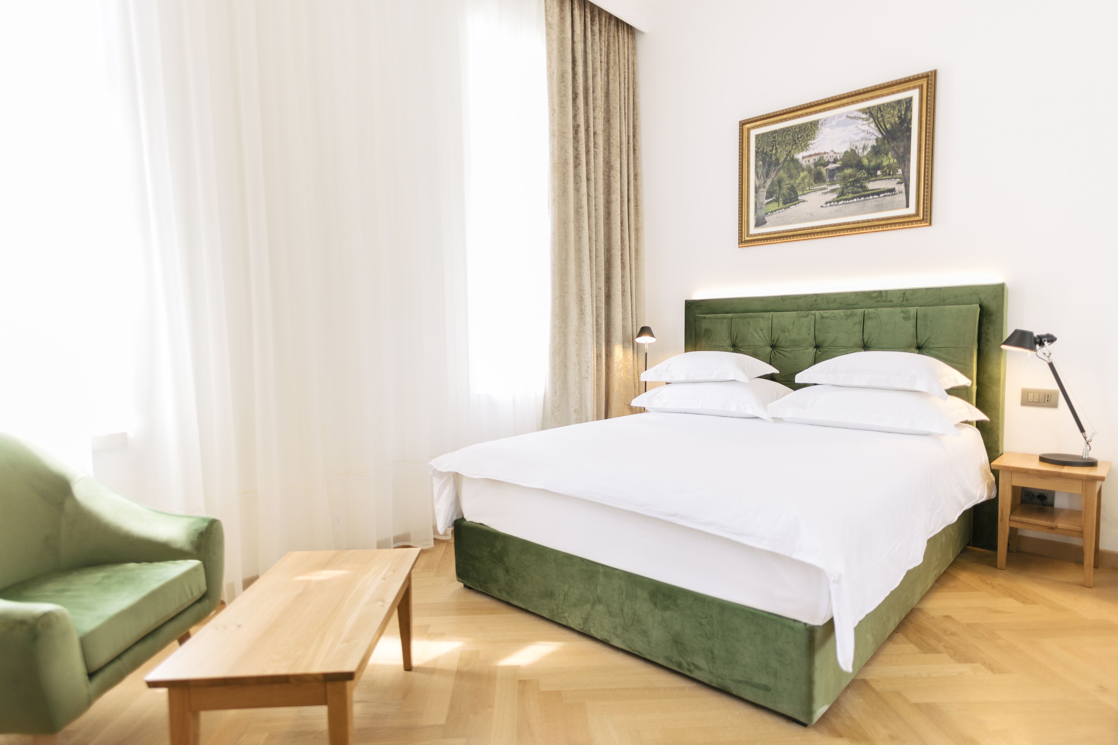 A neatly arranged hotel room with a green upholstered bed, a wooden nightstand, a bedside lamp, a green armchair, and a wooden bench.
