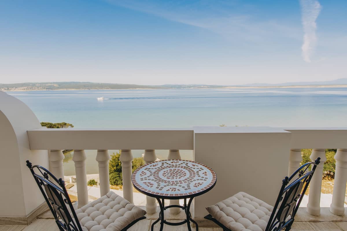 A balcony with a round table and two chairs, overlooking a calm sea and distant hills.