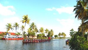 A river with a red boat and a white boat, surrounded by palm trees and a building with a red roof. - Houseboat Cruise in the Backwaters of Kerala (Kottayam)