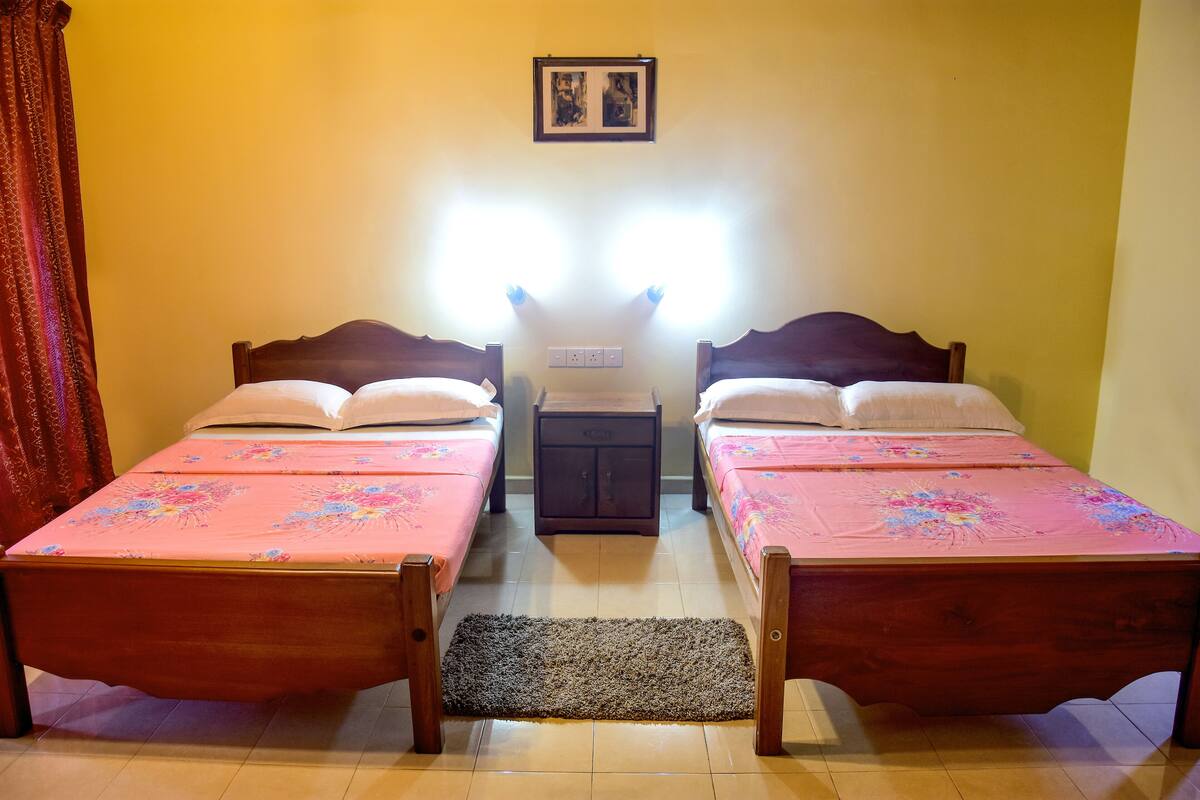 Una habitación con dos camas de madera, cada una con una colcha estampada y almohadas blancas. Entre las camas hay una mesita de madera con un enchufe.