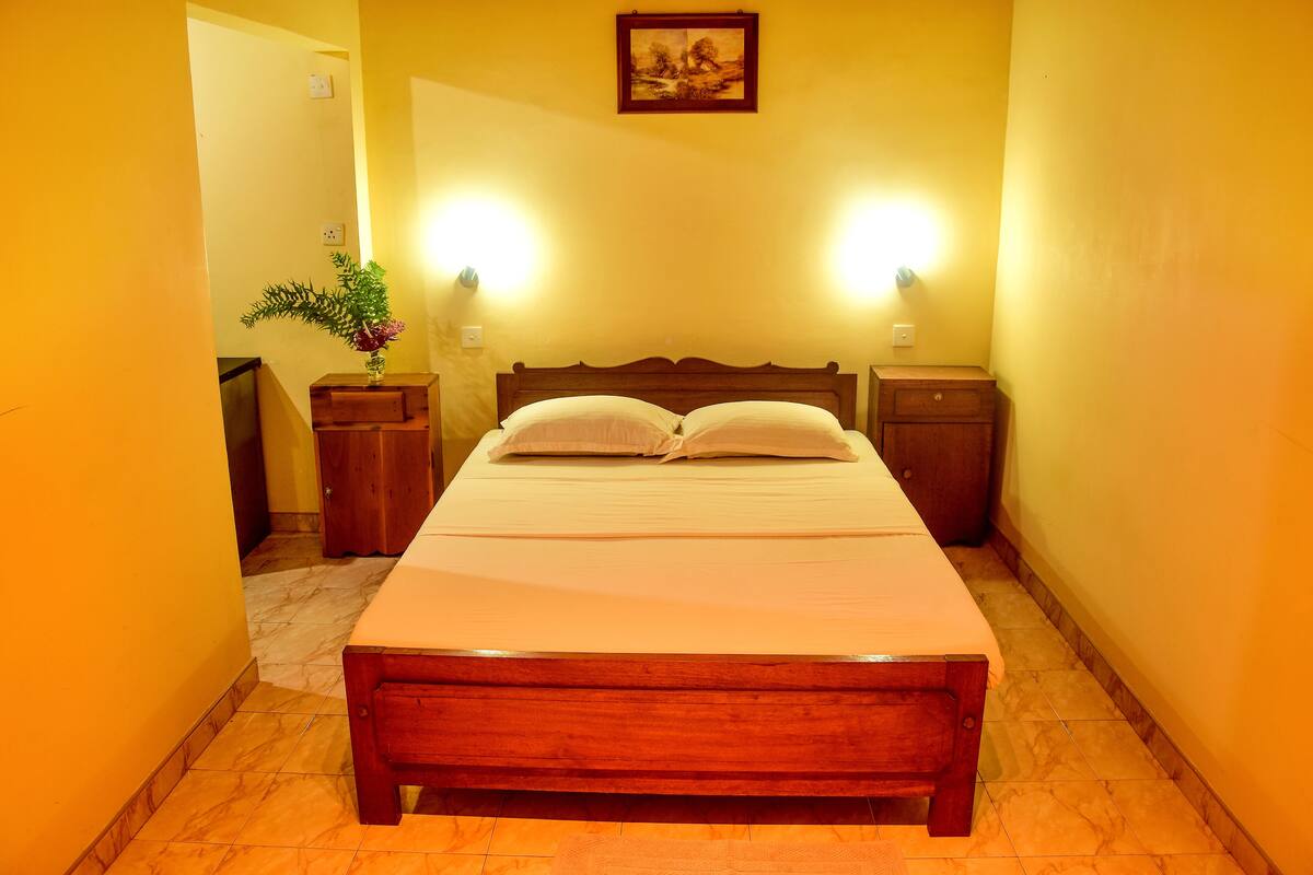 Una cama cuidadosamente tendida en una habitación con paredes amarillas, cabecera de madera y dos mesitas de noche con lámparas.