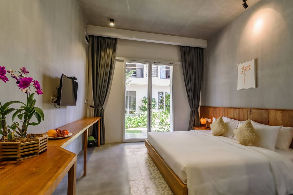 Habitación de hotel con cama, escritorio de madera, planta en maceta, televisor y vistas al exterior.