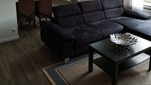 A modern living room with a dark sectional sofa, a dining area with wicker chairs, a kitchen in the background, and a view of the outside through a large window.