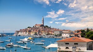 Un pueblo costero con una torre destacada, numerosos barcos en el puerto y una mezcla de edificios tradicionales y modernos.