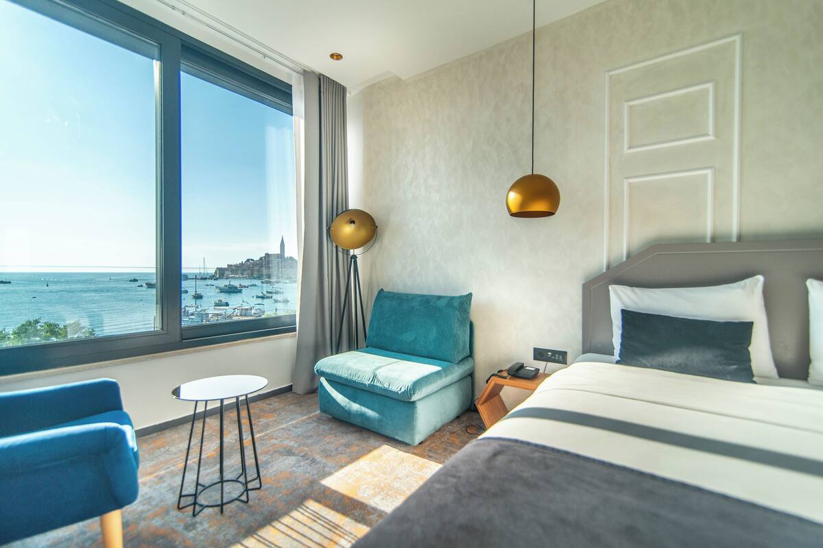 Ein Hotelzimmer mit großem Fenster, einem Bett, einem blauen Sessel, einem kleinen Tisch und einem Nachttisch.