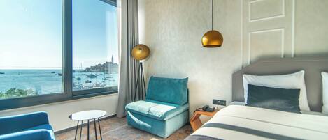 A hotel room with a large window, a bed, a blue chair, a small table, and a nightstand.
