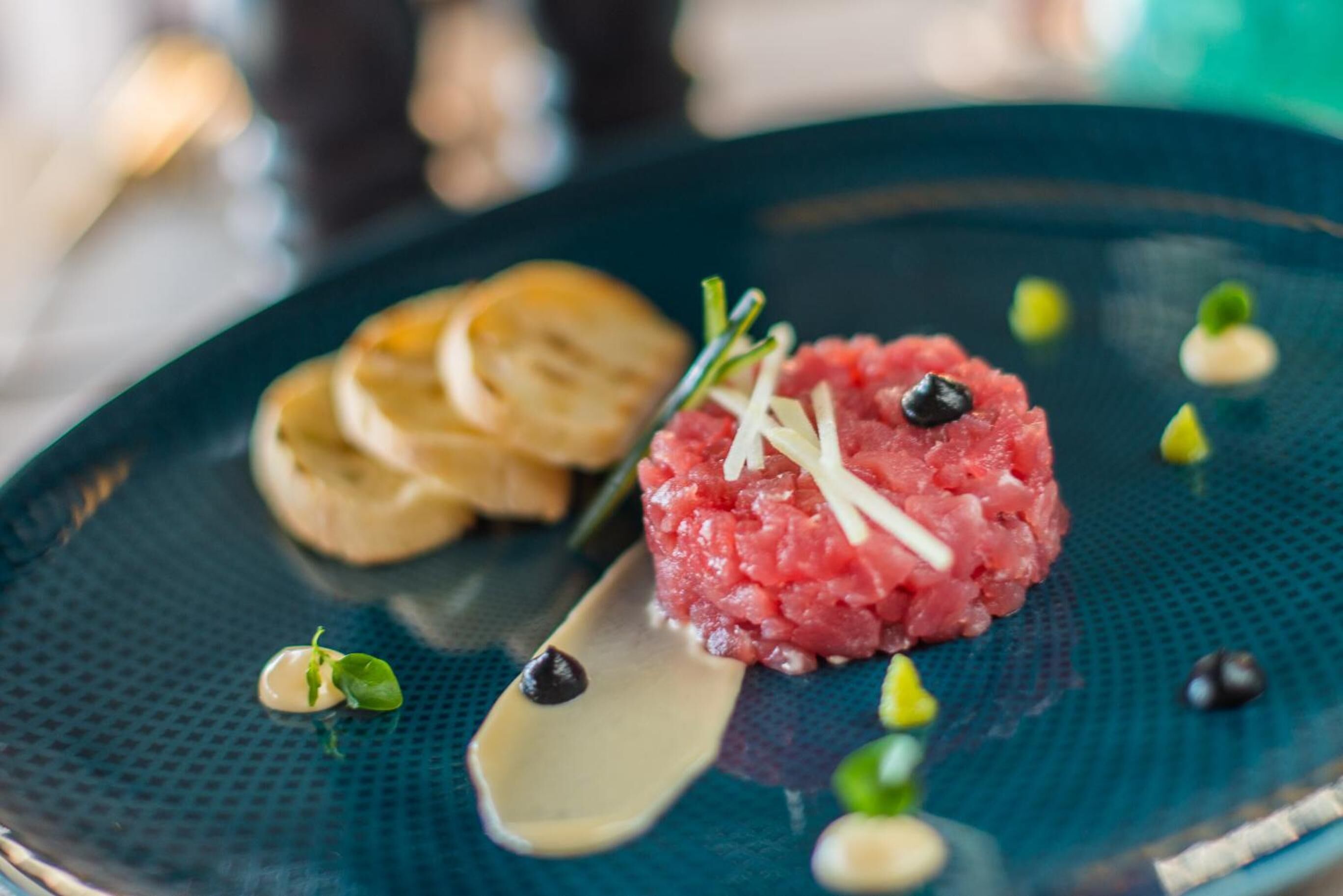 A plate with a dish of raw meat, possibly tartare, garnished with herbs and small green leaves, accompanied by a slice of bread and a small spoon.