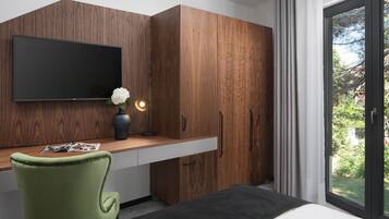 Un dormitorio moderno con un armario de madera, un televisor de pantalla plana, un escritorio con un florero y una revista, una silla verde y un ventanal con vistas a la vegetación.