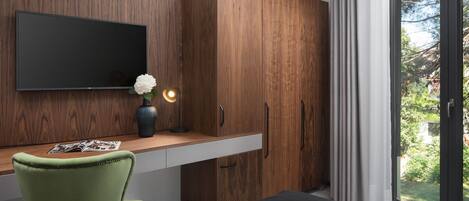Ein modernes Schlafzimmer mit einem Holzschrank, einem Flachbildfernseher, einem Schreibtisch mit Vase und Zeitschrift, einem grünen Sessel und einem großen Fenster mit Blick auf Grünflächen.