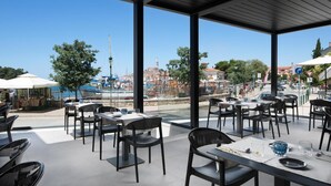 Un área de comedor al aire libre con mesas y sillas, vista a un paisaje urbano y un cuerpo de agua al fondo.