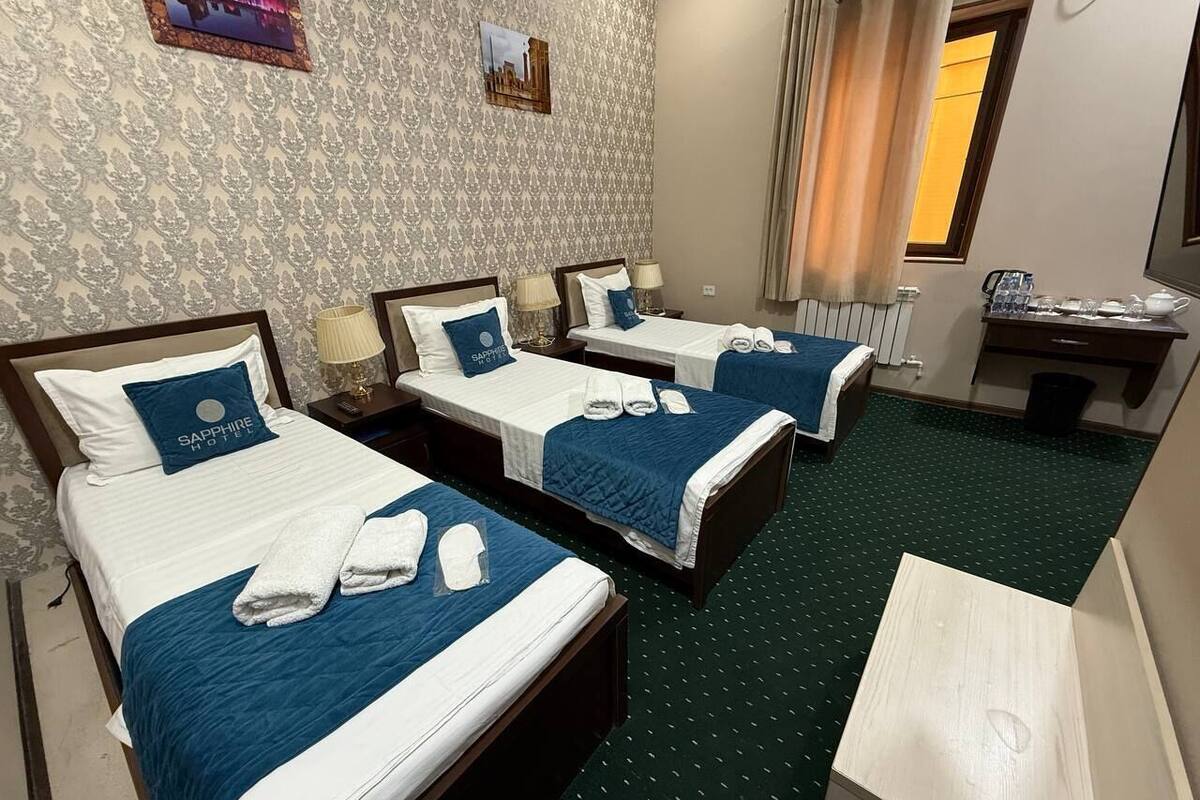 A hotel room with three beds, each with a blue headboard and white bedding. There are towels on each bed, and a small table with a lamp and a coffee maker.