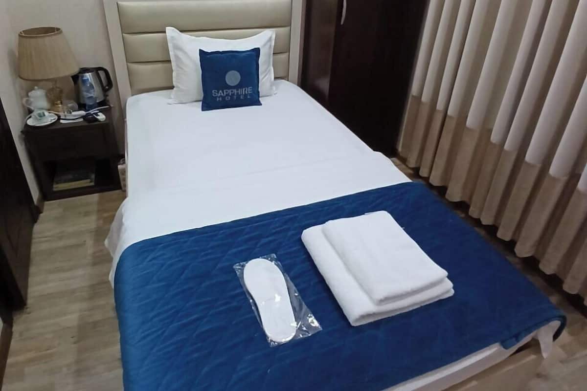 A neatly made bed with a blue bedspread, a white pillow, and a blue pillow with a logo. There is a wooden wardrobe and a nightstand with a lamp and a phone.