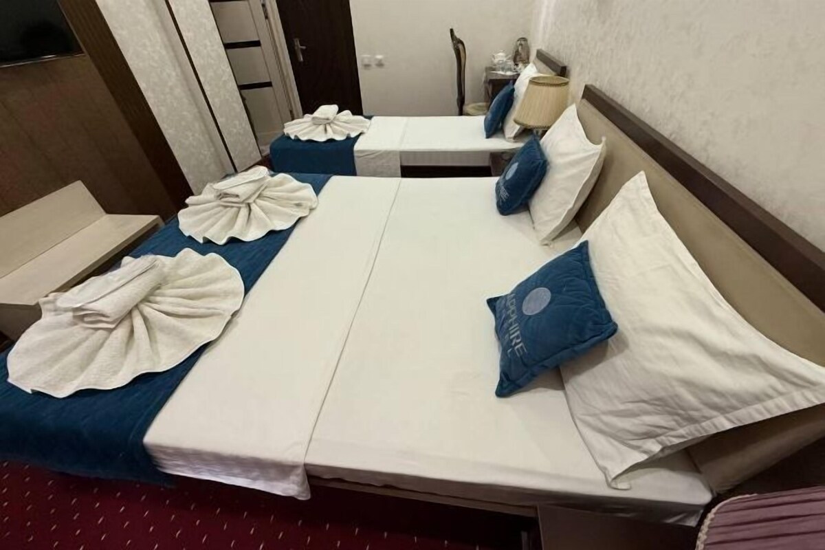 A hotel room with two beds, each with a blue bedspread and white linens. There is a small table with a lamp and a chair.