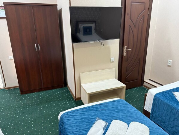 A hotel room with two single beds, a wooden wardrobe, a small wooden bench, and a television mounted on the wall.
