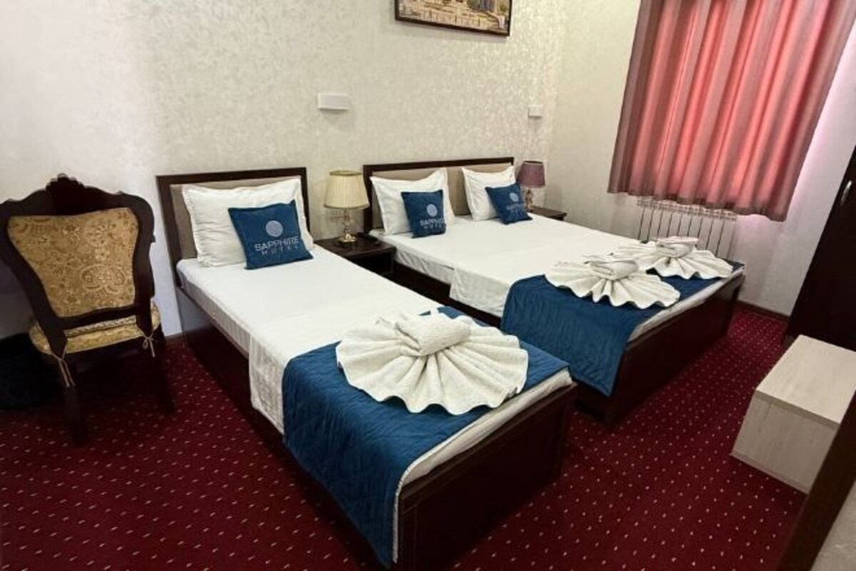 A hotel room with two single beds, each with a blue bedspread and white pillows, a wooden nightstand with a lamp, a chair, and a radiator.