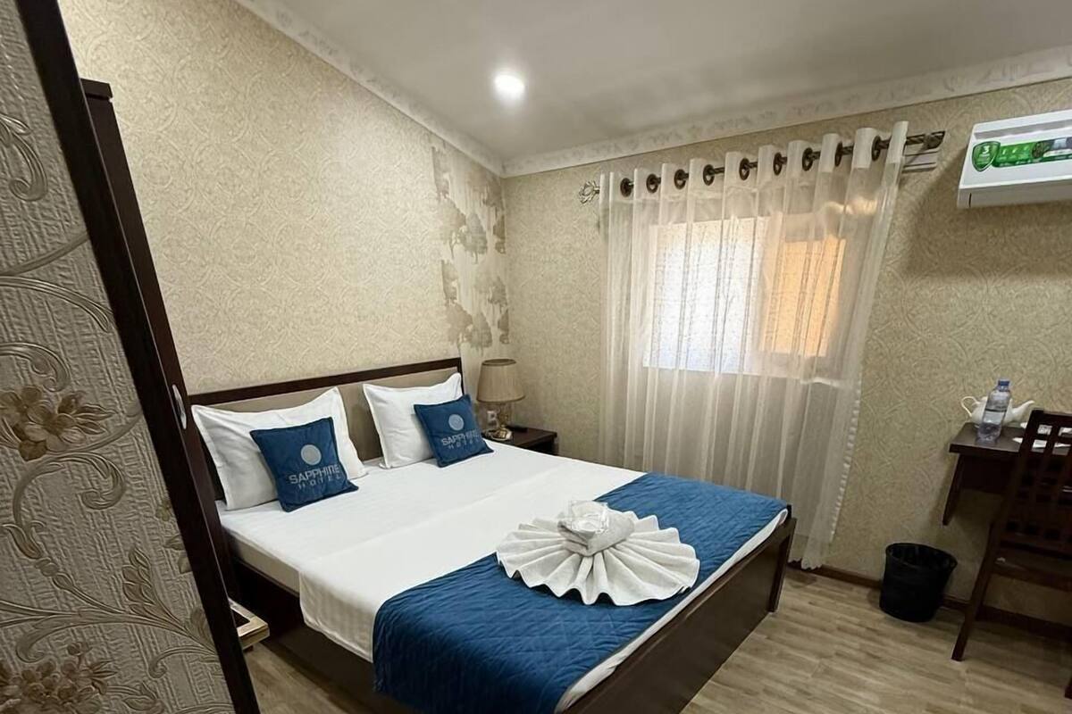 A hotel room with a bed, a desk, a chair, a lamp, and a window with curtains.