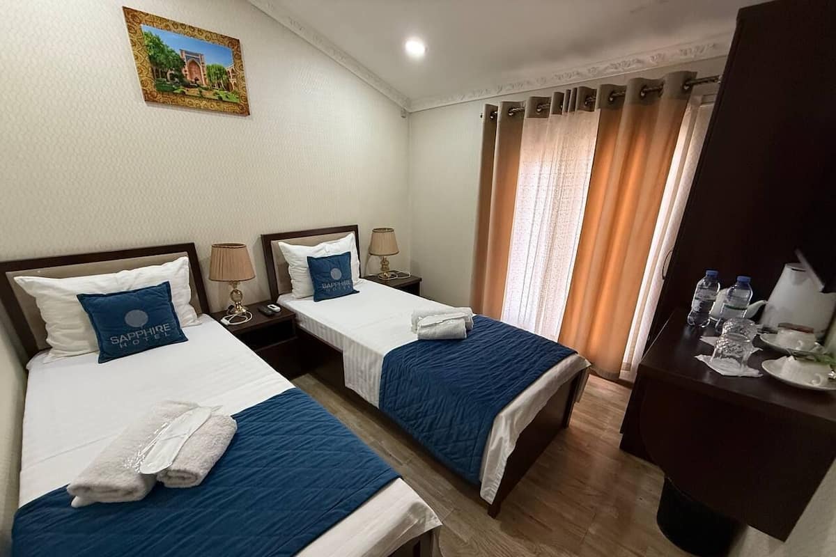 A hotel room with two beds, each with a blue bedspread and matching pillows. There is a small table with a lamp and a picture frame on the wall.
