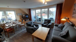 A modern living room with a sofa, coffee table, and dining area. There is a TV on a stand and a window with a view of the outside.