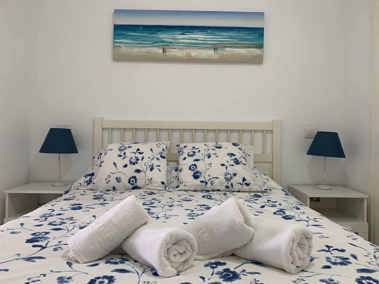 A neatly made bed with floral bedding, two bedside lamps, and a painting of a beach scene above the bed.