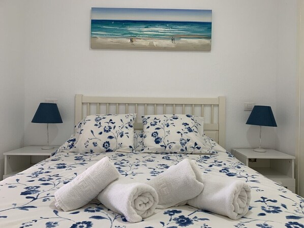 A neatly made bed with floral bedding, two bedside lamps, and a painting of a beach scene above the bed.
