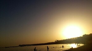 Escena de playa con el sol poniéndose en el horizonte, siluetas de personas y edificios al fondo.