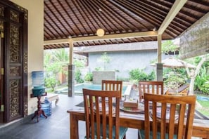 A covered outdoor seating area with wooden furniture and a pool in the background. - Gardenia Gardensbest Breakfast in Bali (Ubud)