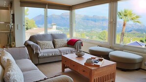 Ein modernes Wohnzimmer mit einer Couch, Hocker und Couchtisch. Große Fenster bieten Blick auf Berge und eine Palme.