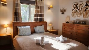 Ein ordentlich bezogenes Bett mit Holzkopfteil, zwei Nachttischlampen, einem Nachttisch, einer Kommode mit Schubladen und einem Fenster mit Vorhängen.