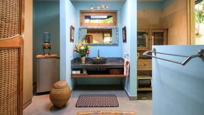 A bathroom with a blue wall, a sink with a mirror above, a small cabinet, a towel rack, and a decorative vase with flowers. - Bungalow Jepunbest Breakfast in Bali (Ubud)