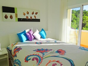 Une chambre avec un lit, des coussins colorés, une table de chevet, une lampe, une fenêtre donnant sur un paysage verdoyant et une œuvre d’art murale.