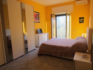 Une chambre à coucher comprenant un lit, un bureau, une chaise, une armoire et une fenêtre donnant sur des immeubles.