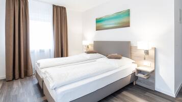 Una cama doble con sábanas blancas, dos mesitas de noche con lámparas, un cuadro en la pared y una ventana con cortinas.