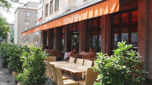 An outdoor seating area with wicker tables and chairs, a red awning with the name "SCHLÜTER'S", and potted plants. - Hotel Köln 2020 (Cologne)