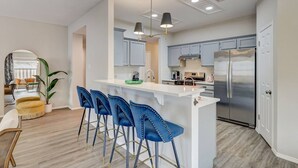 Una cocina moderna con una isla central, taburetes de bar, electrodomésticos de acero inoxidable y un comedor con espejo y sillas.