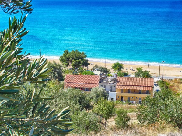 Un bâtiment situé sur la côte, avec un toit recouvert de tuiles rouges, entouré d’oliviers et avec une plage en arrière-plan.