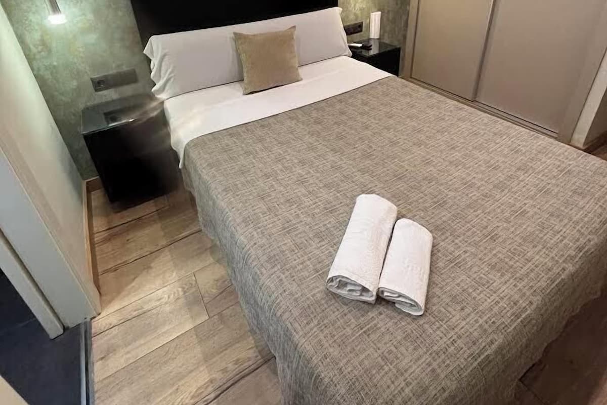 Uma cama bem arrumada com cobertor cinza e toalhas dobradas, mesa de cabeceira com abajur, e luminária fixada na parede.