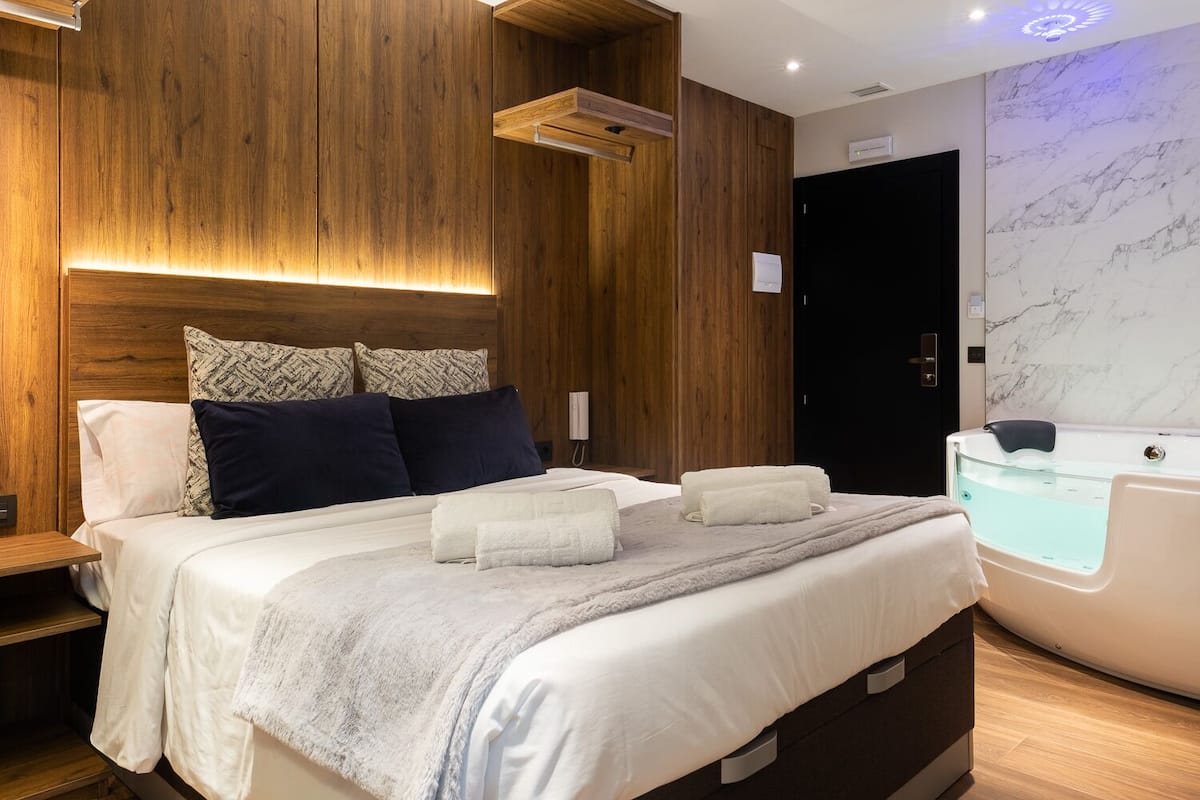 Quarto moderno com cabeceira de madeira, cama grande com várias almofadas, mesa de cabeceira, banheira integrada e parede de mármore.
